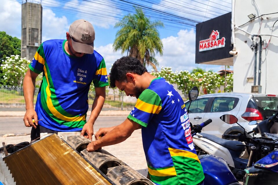 Dois técnicos uniformizados limpando um split piso-teto desmontado em frente ao Supermercado Massapê, em Marituba